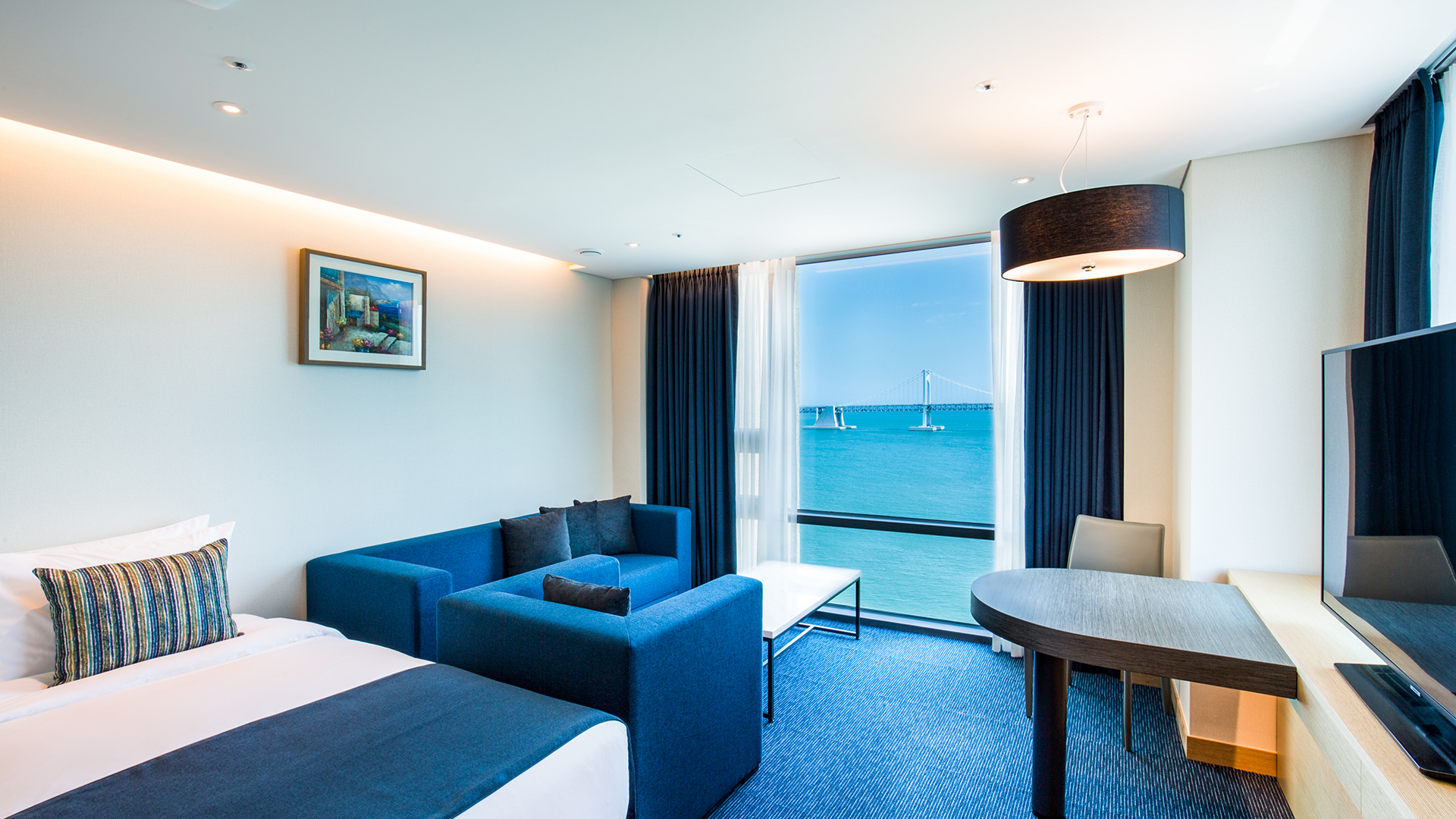 Hotel Central Bay Ocean View 客房真實照片，藍色沙發旁窗外可見廣安里海景與廣安大橋